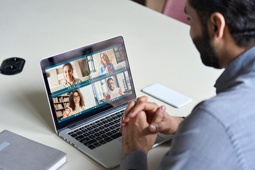 Indian business man having virtual team meeting on video conference call using laptop work from home office talking to diverse people group in remote teamwork online distance chat. Over shoulder view